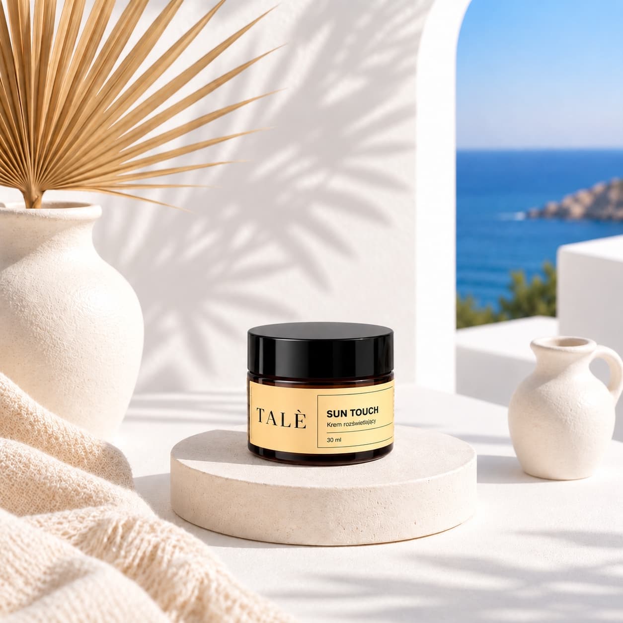 Jar of TALÈ Sun Touch on a stone podium beside a palm leaf, with the sea in the background