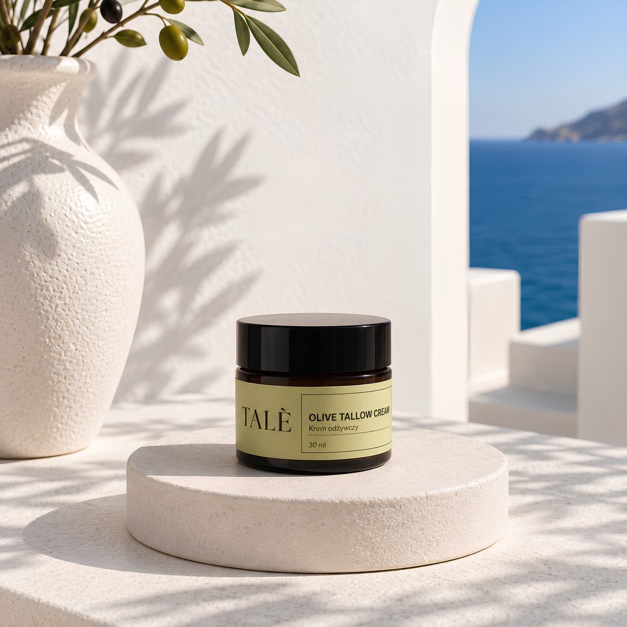 Jar of TALÈ Olive Tallow Cream 30 ml on a white podium beside a vase with an olive branch, with the sea and coastline in the background