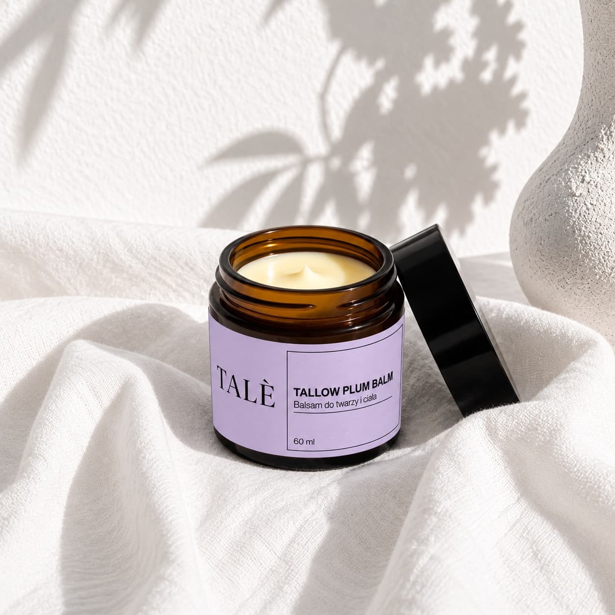 Otwarty słoiczek balsamu TALÈ Tallow Plum Balm na białej tkaninie w cieniach liści